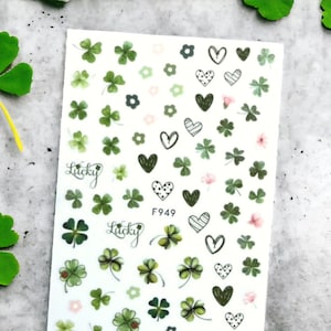 May include: A sheet of nail art stickers with a white background and green four-leaf clover designs, hearts with black and white polka dots, and the word "Lucky" repeated three times. The sheet also includes pink flowers and the code "F949".