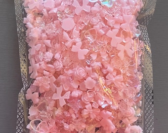 May include: A clear plastic bag filled with an assortment of pink and translucent acrylic embellishments. The bag contains various shapes, including bows, flowers, and other decorative elements, perfect for crafts and DIY projects.