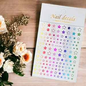 May include: A sheet of rainbow holographic nail decals with a white border and the text "Nail decals - Fashion & Professional -" at the top. The sheet is filled with small, medium, and large star-shaped decals.