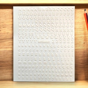 May include: A white sheet of paper with white letters printed on it. The letters are in uppercase and lowercase, and they are arranged in alphabetical order. The letters are in a simple, sans-serif font. The paper is on a wooden surface.