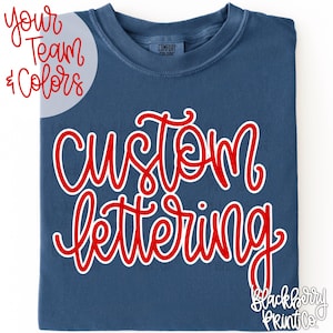 May include: A navy blue crew neck t-shirt featuring the text custom lettering in a red cursive font with a white outline. The shirt has a Comfort Colors label at the inner neckline and includes the text your team & colors in the upper left corner.