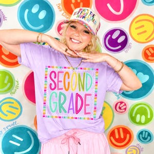 May include: A light purple t-shirt featuring a square graphic with the text "SECOND GRADE" in colorful, multi-colored bubble letters. The shirt is paired with a cream-colored trucker hat with the text "WHAT'S THE BEST THAT COULD HAPPEN?" and pink shorts.