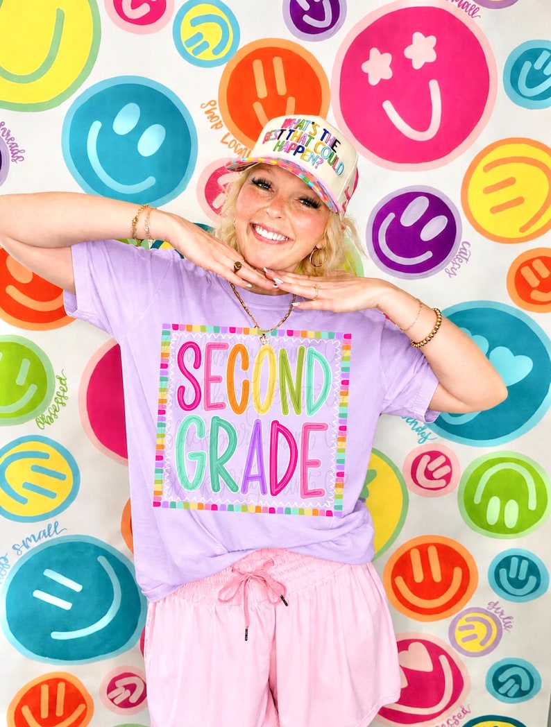 May include: A light purple t-shirt featuring a square graphic with the text "SECOND GRADE" in colorful, multi-colored bubble letters. The shirt is paired with a cream-colored trucker hat with the text "WHAT'S THE BEST THAT COULD HAPPEN?" and pink shorts.