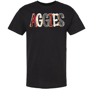May include: Black t-shirt featuring the word "AGGIES" in a distressed, collegiate font. The letters are in red, white, grey, and gold. The shirt has a crew neck and short sleeves.