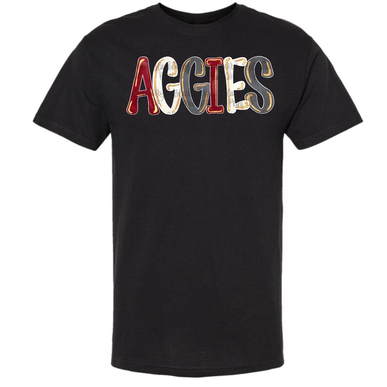 May include: Black t-shirt featuring the word "AGGIES" in a distressed, collegiate font. The letters are in red, white, grey, and gold. The shirt has a crew neck and short sleeves.