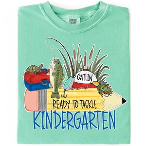 May include: A mint green t-shirt featuring a graphic of a yellow pencil, a red and blue tackle box, a turtle, a fishing rod, and a fish. The text reads GATLIN, READY TO TACKLE, and KINDERGARTEN in black and blue lettering across the center design.