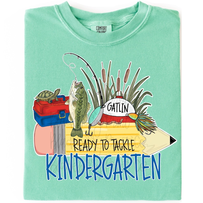 May include: A mint green t-shirt featuring a graphic of a yellow pencil, a red and blue tackle box, a turtle, a fishing rod, and a fish. The text reads GATLIN, READY TO TACKLE, and KINDERGARTEN in black and blue lettering across the center design.