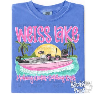 May include: A periwinkle blue t-shirt featuring a graphic of a pink bass boat on water with palm trees and a yellow sun. The text reads WEISS LAKE in pink at the top and Making Waves + Catching Rays in pink cursive script at the bottom.