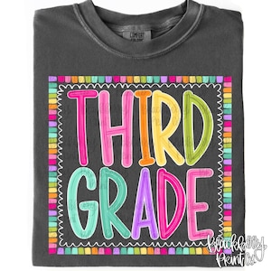 May include: A charcoal gray t-shirt featuring the text "THIRD GRADE" in large, colorful, outlined letters. The design is enclosed in a scalloped square border made of small, multicolored rectangular blocks in shades of pink, orange, yellow, green, and blue.
