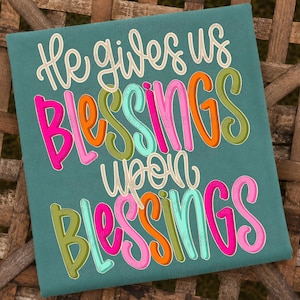 May include: A teal t-shirt featuring the text "He gives us Blessings upon Blessings" in a multi-coloured script font. The words are written in shades of pink, orange, olive green, and light blue, with a white outline on a solid teal background.