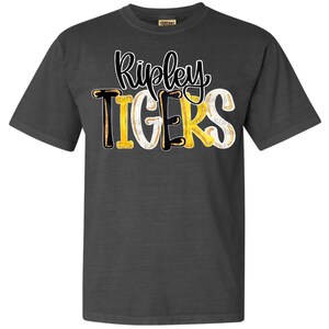 May include: Dark grey t-shirt featuring the text "Ripley TIGERS". The word "Ripley" is in a cursive font, and the word "TIGERS" is in a mix of black, yellow, and white lettering. The shirt is made of a soft material.