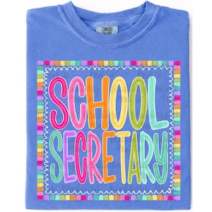 May include: A periwinkle blue t-shirt featuring a colorful graphic print with the text SCHOOL SECRETARY in bright pink, orange, yellow, and green letters. The text is enclosed within a decorative border made of small, multicolored rectangular blocks.