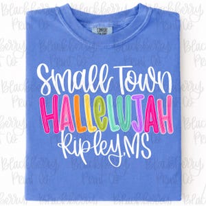 Puede incluir: Una camiseta azul con las palabras "Small Town Hallelujah Ripley, MS" en blanco y letras coloridas. La camiseta es de un material suave y tiene cuello redondo.