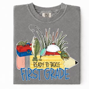 May include: A grey t-shirt featuring a graphic of a yellow pencil, a green fish, a turtle on a red and blue tackle box, a fishing rod, and cattails. The text reads "READY TO TACKLE FIRST GRADE" in black and blue lettering on the pencil design.