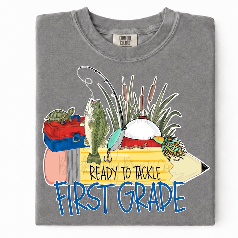 May include: A grey t-shirt featuring a graphic of a yellow pencil, a green fish, a turtle on a red and blue tackle box, a fishing rod, and cattails. The text reads "READY TO TACKLE FIRST GRADE" in black and blue lettering on the pencil design.