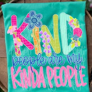 May include: A teal t-shirt featuring the word KIND in large, multi-colored letters decorated with floral illustrations. Below, the text reads people are my KINDA PEOPLE in blue cursive and bold pink block letters with a white outline.