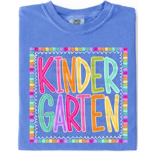May include: A periwinkle blue crew neck t-shirt featuring a colorful graphic print. The design displays the word KINDERGARTEN in multi-colored, bubble-style letters surrounded by a scalloped border made of small, vibrant, multi-colored squares.