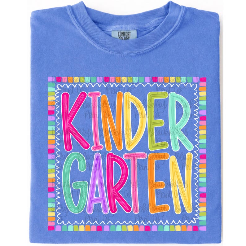 May include: A periwinkle blue crew neck t-shirt featuring a colorful graphic print. The design displays the word KINDERGARTEN in multi-colored, bubble-style letters surrounded by a scalloped border made of small, vibrant, multi-colored squares.
