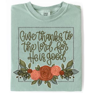 May include: A sage green t-shirt featuring the text "Give thanks to the Lord for He is good" in dark green cursive script. Below the text is a floral illustration with red and peach roses, green leaves, and dark berries, all framed by a dotted border.
