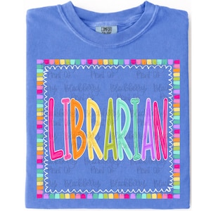 May include: A periwinkle blue t-shirt featuring the word LIBRARIAN in colourful, outlined letters. The text is enclosed in a white scalloped border surrounded by a frame of small, multicoloured rectangular blocks. The shirt has a crew neckline.