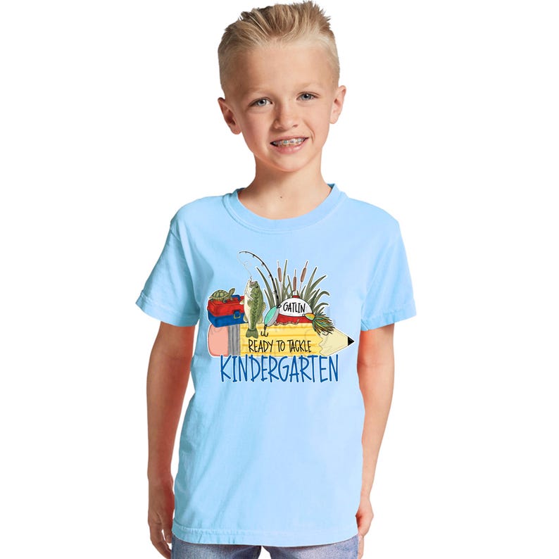 May include: A light blue t-shirt featuring a graphic of a yellow pencil with fishing gear, including a tackle box, a fish, a bobber, and reeds. The text reads "READY TO TACKLE" and "KINDERGARTEN" in blue, with the name "GATLIN" on the bobber.