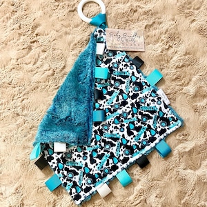 May include: A square baby security blanket featuring a white fabric with a black cow print and turquoise text, backed with textured teal plush fabric. It includes multiple turquoise, black, and white ribbon loops and a white plastic ring. A tag reads Baby Bundles by Maddy.