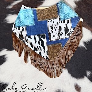 May include: A triangular bandana featuring a patchwork design of blue floral fabric, brown paisley print, and black and white cowhide pattern. The bottom edge is finished with long brown fringe. The text Baby Bundles by Maddy HANDMADE is visible.