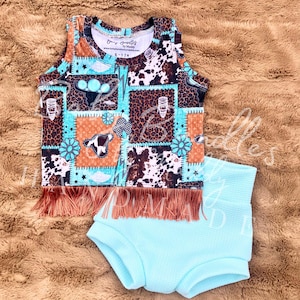 May include: A two-piece baby outfit featuring a patchwork tank top and high-waisted shorts. The tank top has a brown fringe and a mix of patterns, including cow print and turquoise accents. The shorts are a solid light blue color.