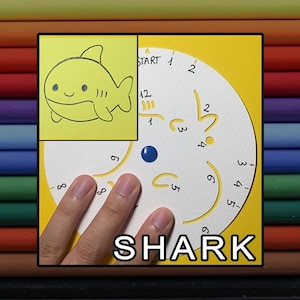 May include: A white circular drawing stencil with cutouts for creating a cartoon shark, featuring numbers and lines for guidance. The word SHARK is printed at the bottom. A yellow inset shows the finished shark drawing with a smiling face.