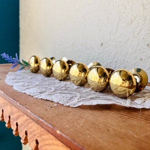 May include: A row of eight gold-toned cabinet knobs with a round shape and a smooth, shiny finish. The knobs are arranged in a line on a white lace doily.