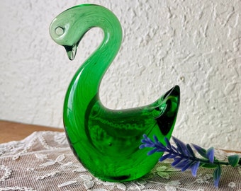 May include: A green glass swan figurine with a smooth, rounded body and a long, curved neck. The swan is sitting on a white lace tablecloth.