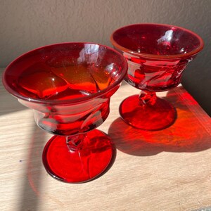 May include: Two red glass stemmed cups with a textured design. The cups are empty and sitting on a wooden surface.