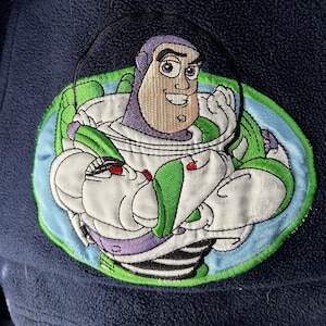 May include: A blue fleece jacket featuring an embroidered image of Buzz Lightyear from the Toy Story franchise. The image depicts Buzz in his space ranger suit, helmet off, holding his laser gun. The text "STAR" is embroidered on the left side of the jacket and "WAR" is embroidered on the right side.