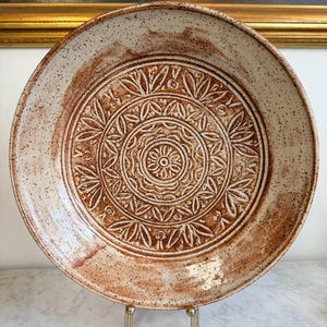 May include: A large, round ceramic plate with a detailed, embossed floral pattern in the center. The plate has a speckled, off-white and brown color scheme. The rim is slightly raised, and the plate is displayed on a gold-colored stand.