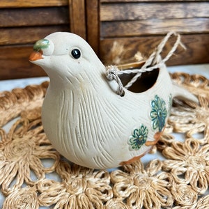 May include: A white ceramic bird planter with a brown, green, and blue floral design. The planter has a hole in the back for hanging and a rope loop.