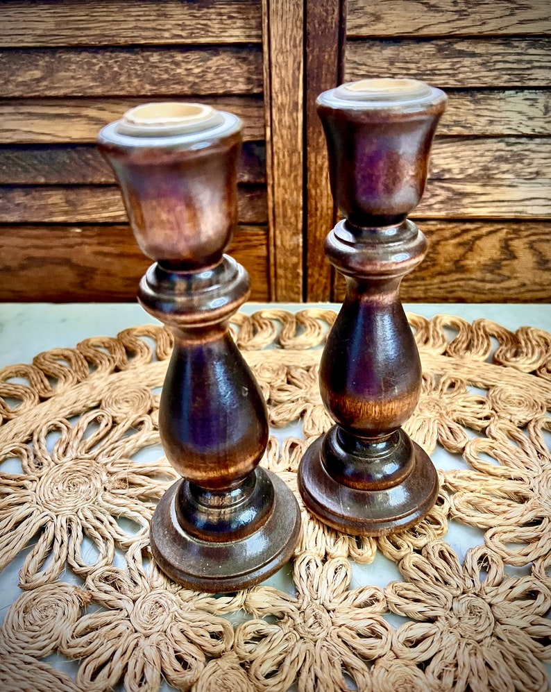 May include: Two wooden candle holders with a dark brown finish. The candle holders are on a woven straw placemat.