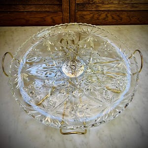 May include: A clear glass serving tray with a starburst pattern and gold-colored metal handles and support. The tray is oval-shaped and features a central decorative element. The tray is designed for serving food or displaying items.