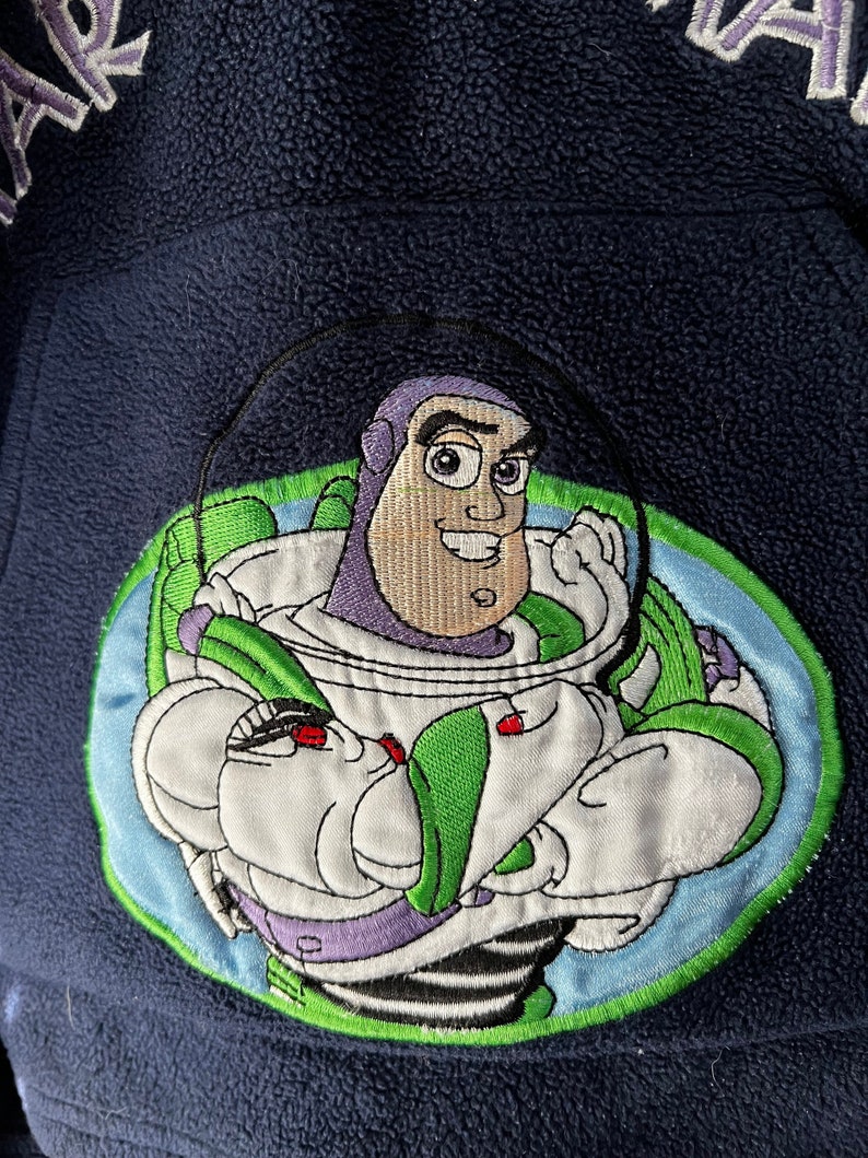 May include: A blue fleece jacket featuring an embroidered image of Buzz Lightyear from the Toy Story franchise. The image depicts Buzz in his space ranger suit, helmet off, holding his laser gun. The text "STAR" is embroidered on the left side of the jacket and "WAR" is embroidered on the right side.