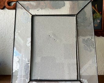 May include: A silver picture frame with a floral design. The frame has a clear glass front and a silver metal frame. The floral design is in a white and gray color scheme.