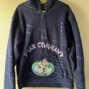 May include: A navy blue fleece hoodie with a zip fastening and a hood. The hoodie features a large embroidered patch of Buzz Lightyear from the Toy Story franchise with the text "Star Command" above it.