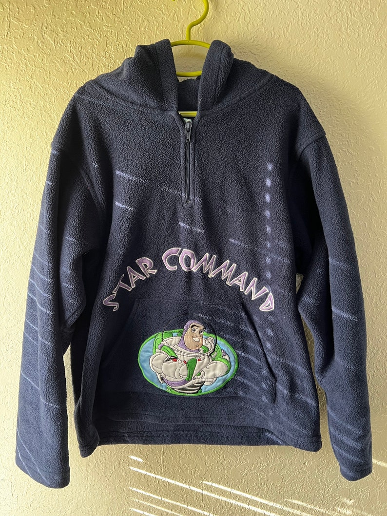 May include: A navy blue fleece hoodie with a zip fastening and a hood. The hoodie features a large embroidered patch of Buzz Lightyear from the Toy Story franchise with the text "Star Command" above it.