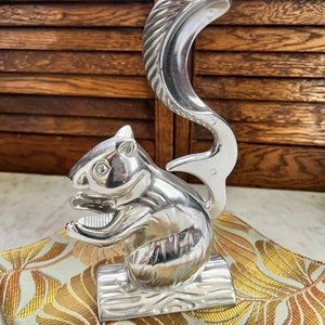 May include: A silver squirrel nutcracker with a detailed, textured tail. The nutcracker is sitting on a patterned fabric with gold and green leaves.