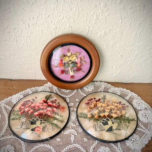 May include: Three vintage floral art pieces. One round wooden framed piece with a pink background and a cross with flowers. Two oval pieces with dried flower arrangements in vases, set on a white lace doily.