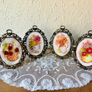 May include: Four oval-shaped framed floral embroidery pieces with a white background and colorful flowers. The frames are gold metal with ornate designs.