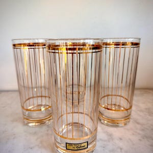 May include: Four clear highball glasses with gold vertical stripes and gold rims. The glasses have a rectangular label that reads "22K GOLD by Culver". The glasses are on a white marble surface.