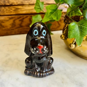 May include: A small, black ceramic figurine of a dog sitting with its head tilted down. The dog has white spots on its face and a red nose. The dog's eyes are blue.