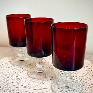 May include: Three ruby red wine glasses with clear stems and bases. The glasses have a cylindrical shape and are arranged in a row. The glasses are on a cream-colored, crocheted doily.