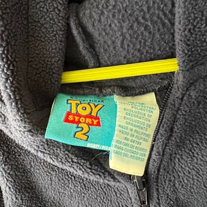 May include: A black fleece zip-up hoodie with a Toy Story 2 label. The label is blue and yellow with the words "Toy Story 2" in white. The label also includes information about the fabric content and care instructions.