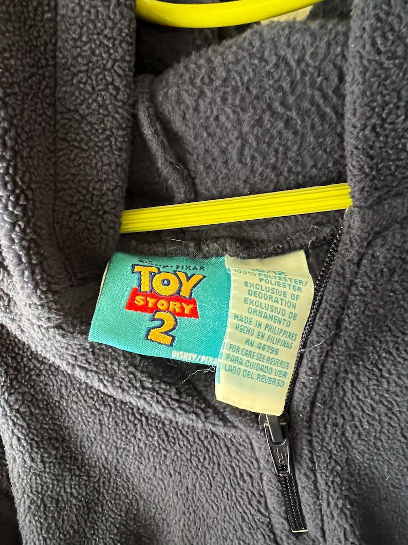 May include: A black fleece zip-up hoodie with a Toy Story 2 label. The label is blue and yellow with the words "Toy Story 2" in white. The label also includes information about the fabric content and care instructions.