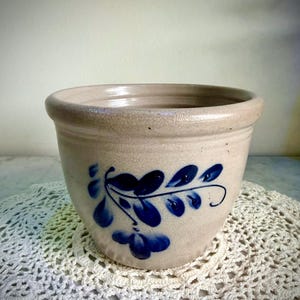 May include: A beige stoneware planter with a blue floral design. The planter has a rounded shape with a slightly flared rim. The blue design features stylized leaves and stems. The planter is sitting on a white crocheted doily.